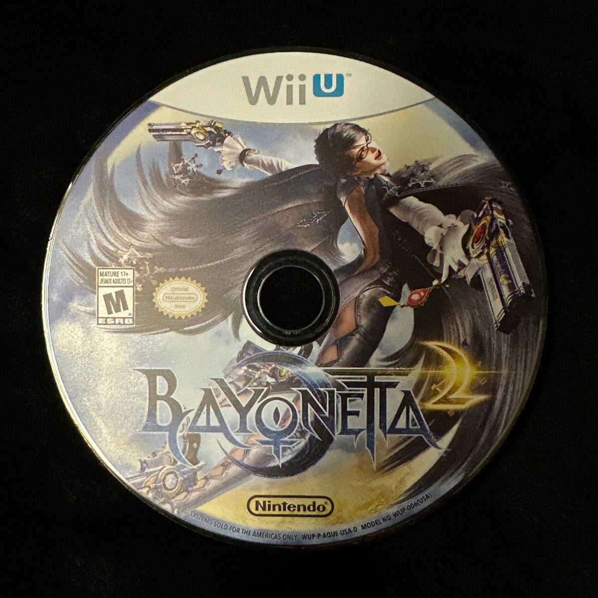 A Wii U game disc for Bayoneta 2 on Nintendo Wii, featuring artwork of a female warrior with a weapon, resting on a black fabric surface.