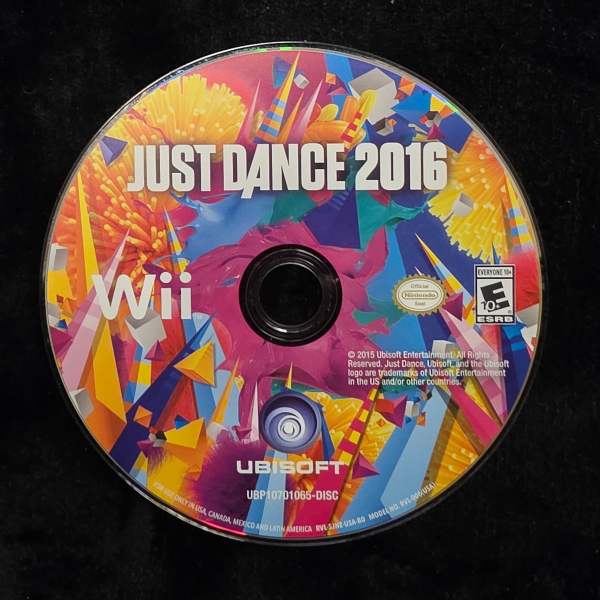 A used Nintendo Wii game disc for Just Dance 2016, featuring a vibrant multicolor abstract cover design, resting on a black fabric surface.