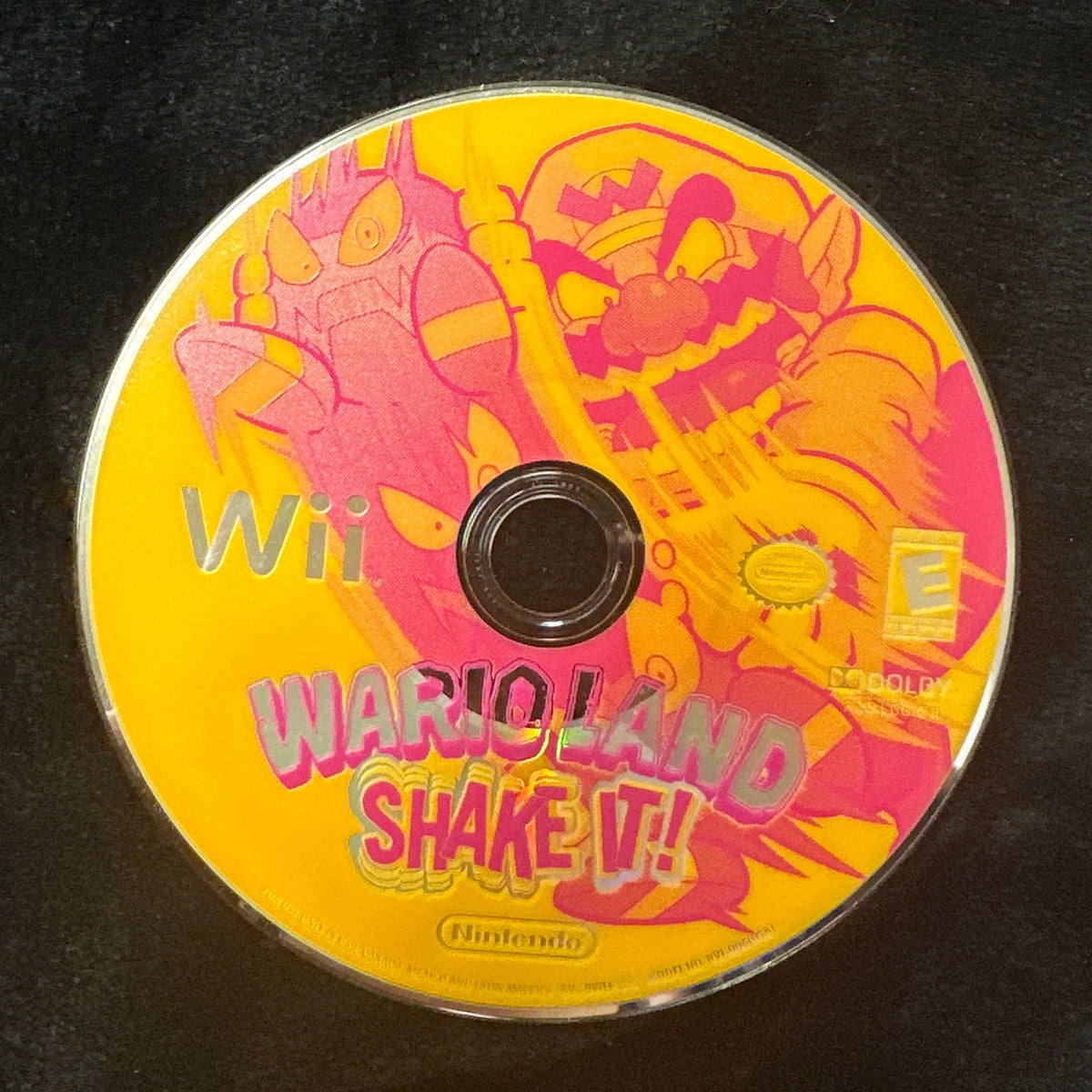 A Wii game disc for 'Wario Land Shake It!' resting on a black fabric surface, with a dark tiled floor visible in the background.