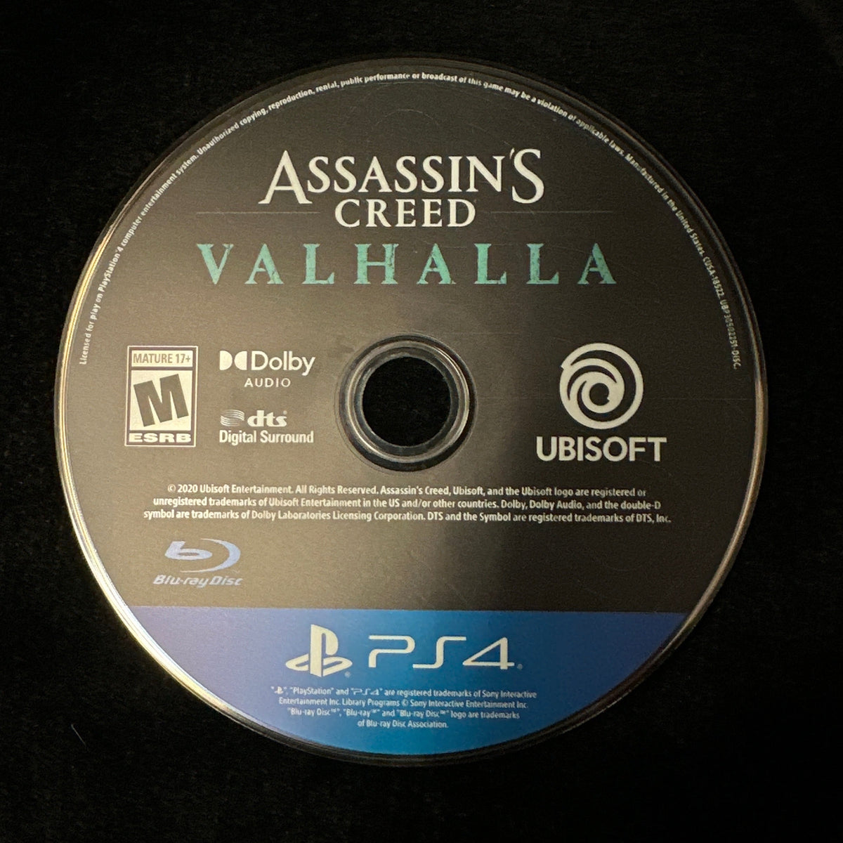 A PlayStation 4 game disc for Assassin's Creed Valhalla, featuring the Ubisoft and Sony PlayStation branding, photographed on a black background.