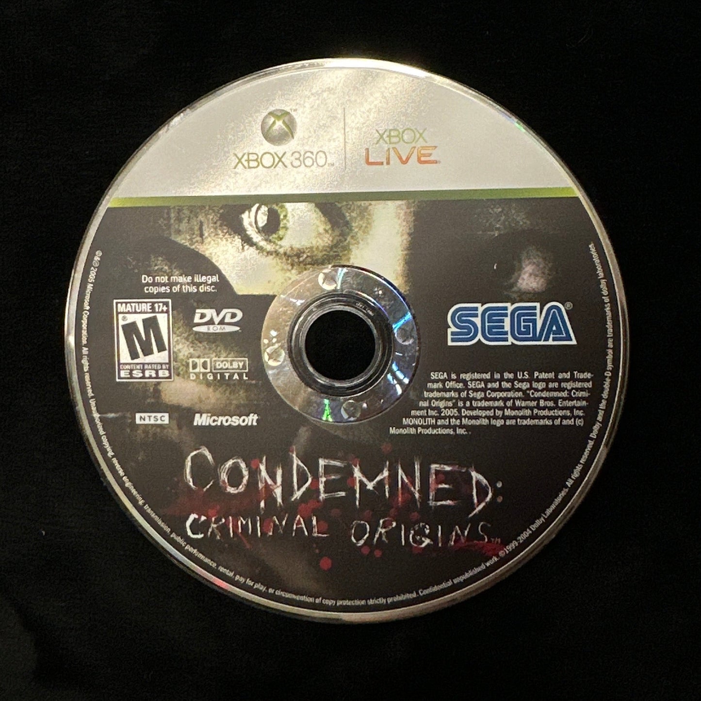 A used Xbox 360 game disc for 'Condemned: Criminal Origins' resting on a black fabric surface. The disc shows the Xbox 360 logo, Xbox Live branding, the SEGA logo, and the Microsoft logo.