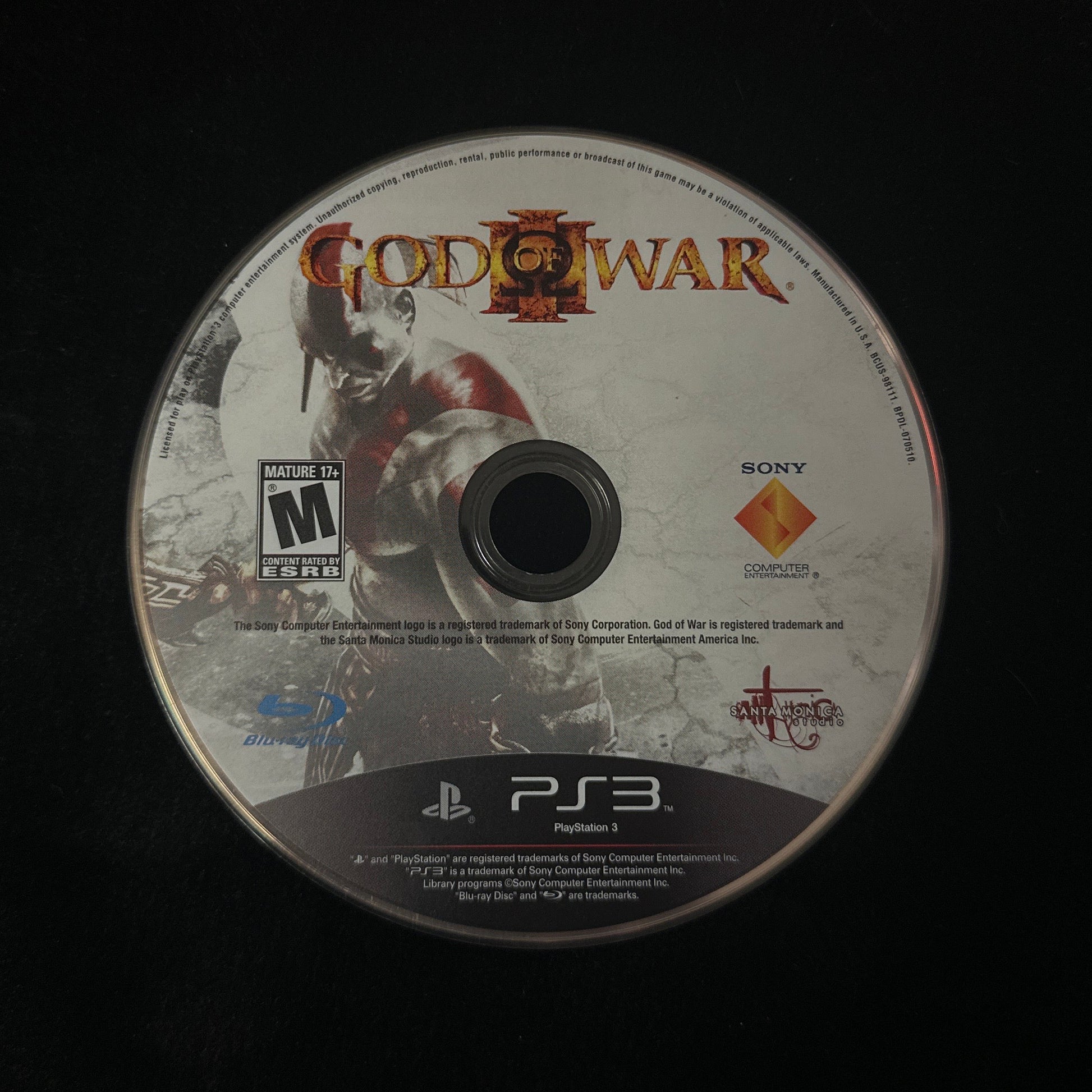 A used PlayStation 3 God of War II game disc resting on a black surface. The disc features the game's cover art, the God of War logo, an M rating from ESRB, and PlayStation 3 and Sony branding.