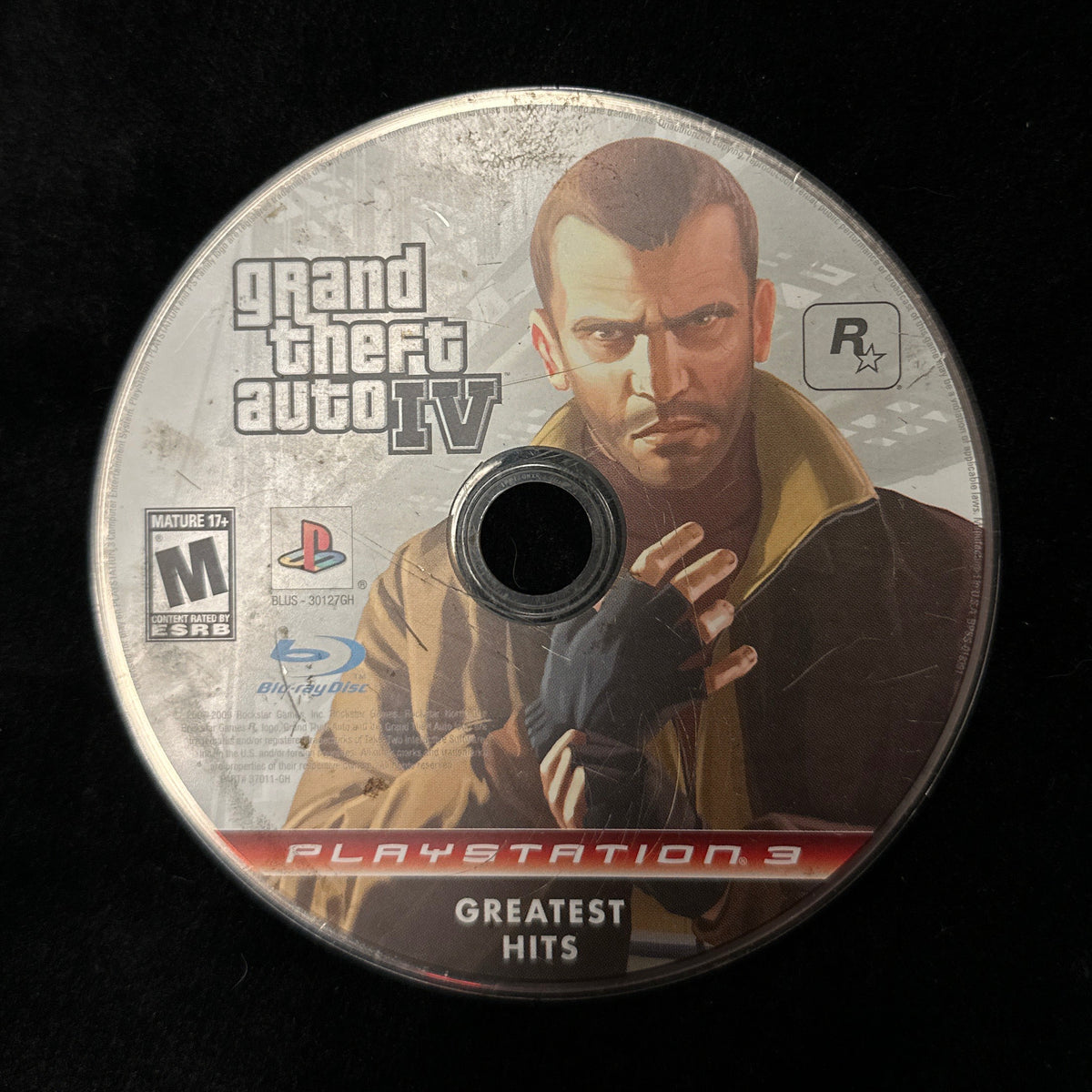 A PlayStation 3 'Grand Theft Auto IV Greatest Hits' game disc resting on a black fabric surface.