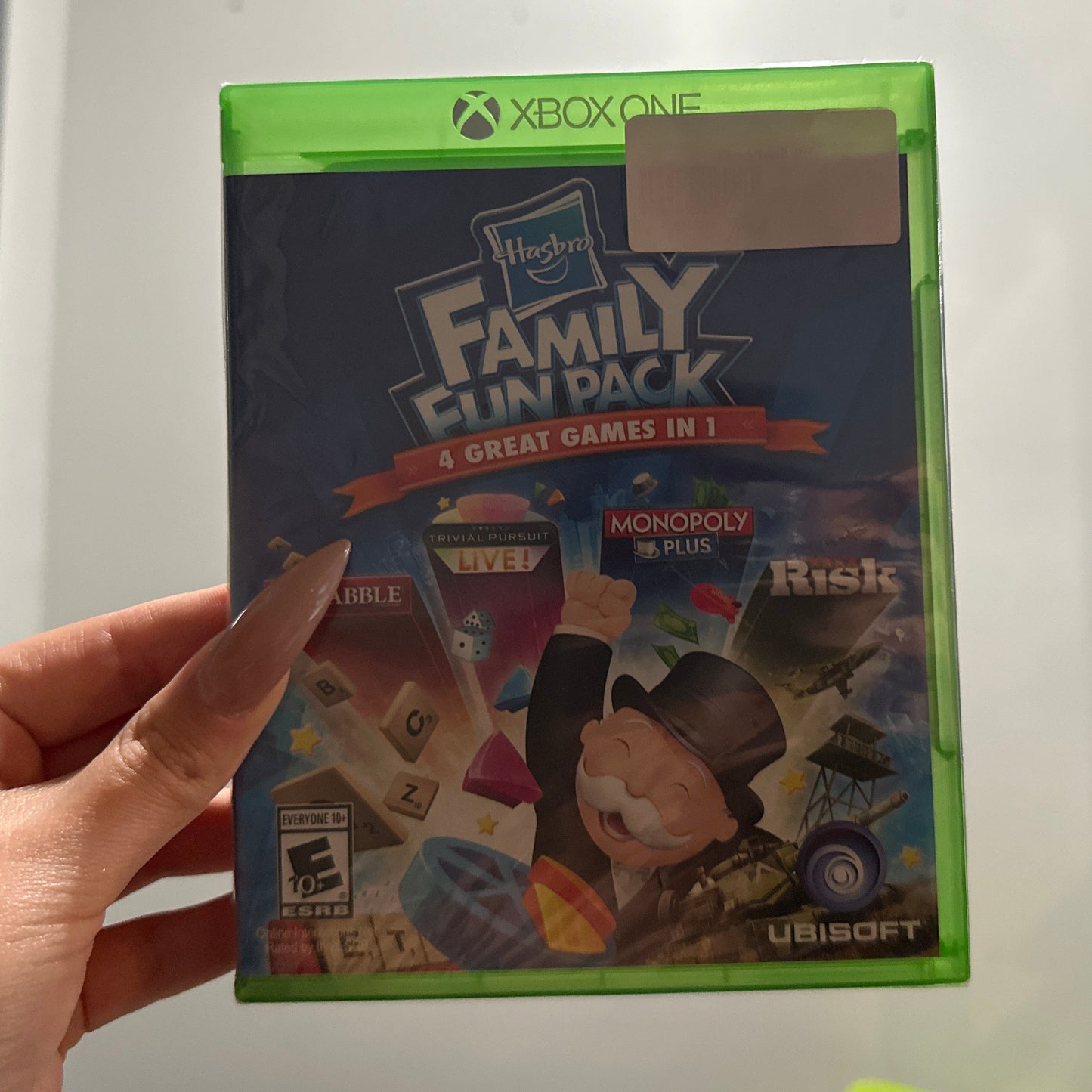 Hand holding a 'Family Fun Pack' video game for Xbox One against a white wall.