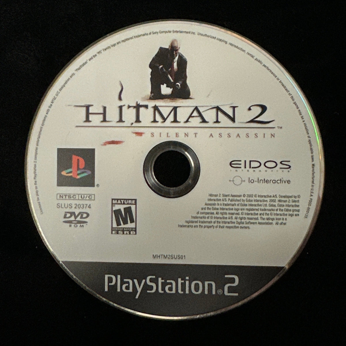 A PlayStation 2 game disc for Hitman 2, resting on a black fabric surface. The disc features the PlayStation 2 logo, the Hitman 2 title, and the Eidos brand name.