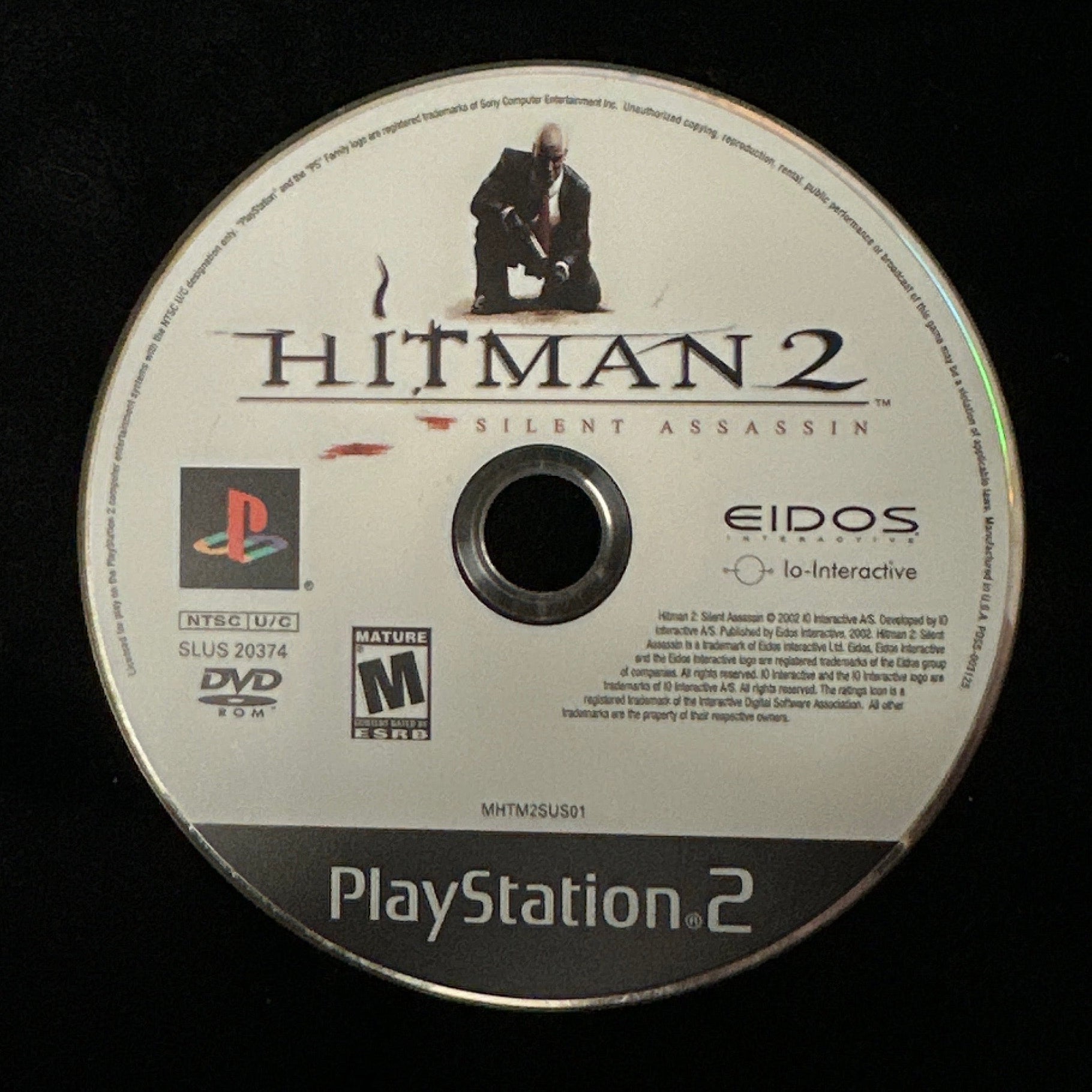 A PlayStation 2 game disc for Hitman 2, resting on a black fabric surface. The disc features the PlayStation 2 logo, the Hitman 2 title, and the Eidos brand name.
