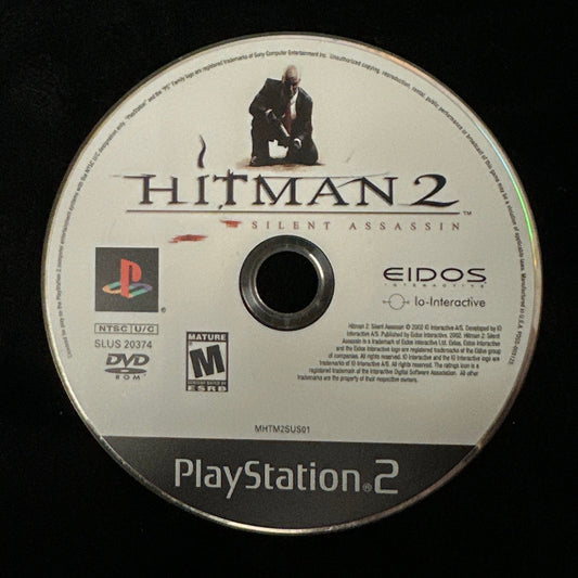 A PlayStation 2 game disc for Hitman 2, resting on a black fabric surface. The disc features the PlayStation 2 logo, the Hitman 2 title, and the Eidos brand name.