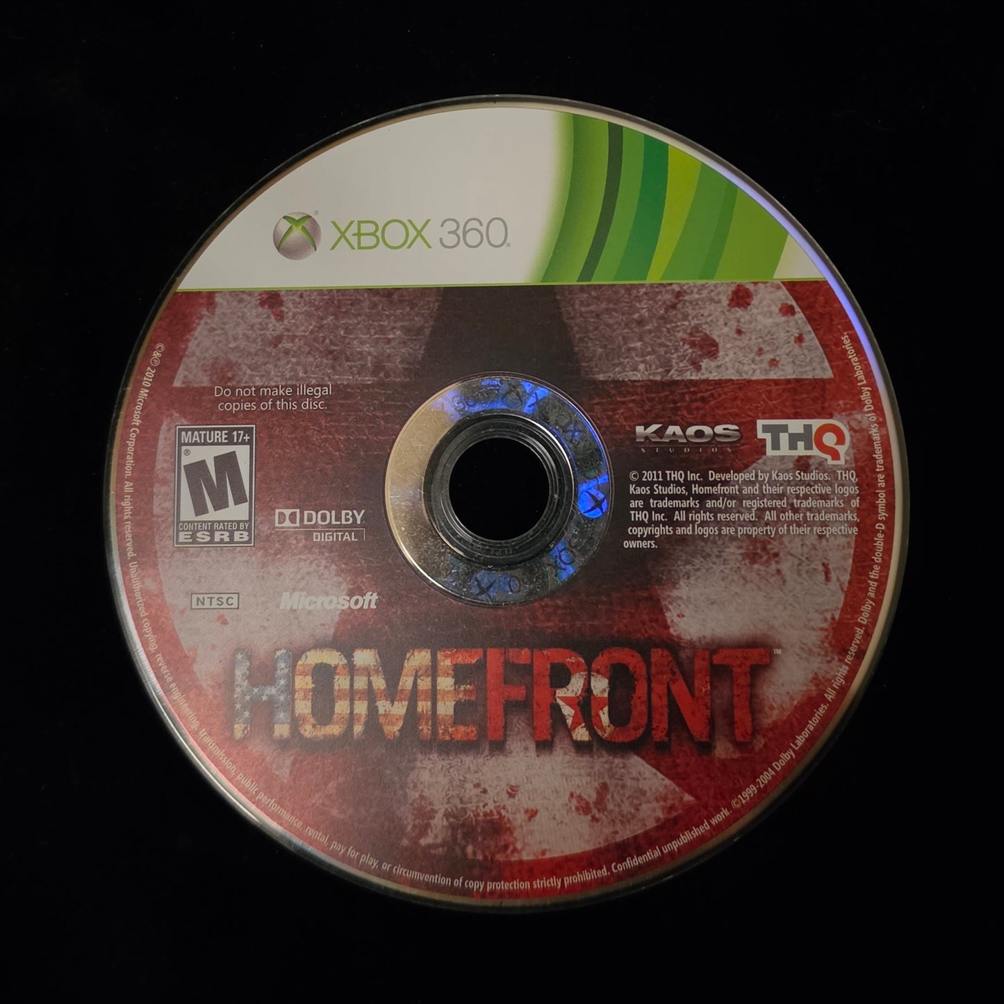 An Xbox 360 game disc for the video game Homefront, featuring a red and white splatter design with green and white Xbox branding, resting on a black surface.