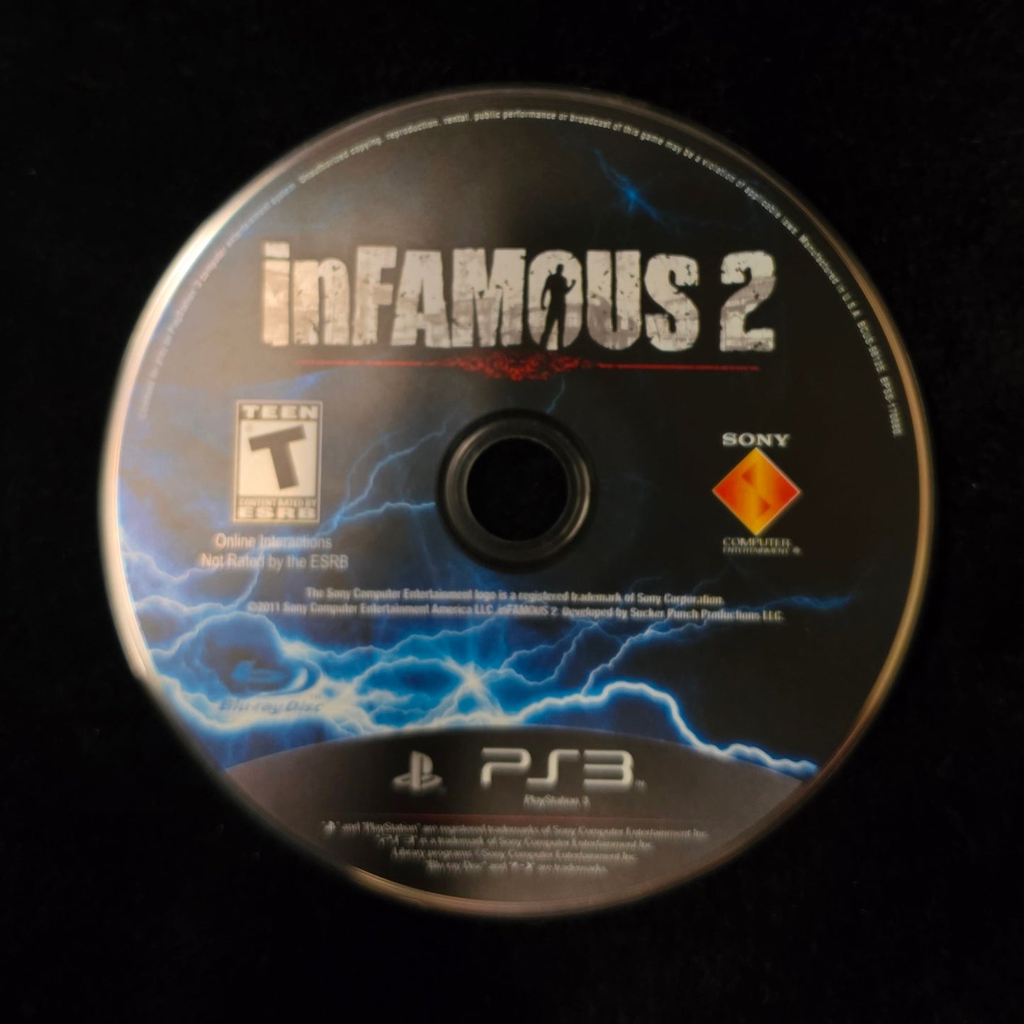 Infamous 2 PlayStation 3 game disc resting on a black surface, showing the PlayStation logo and PS3 branding.