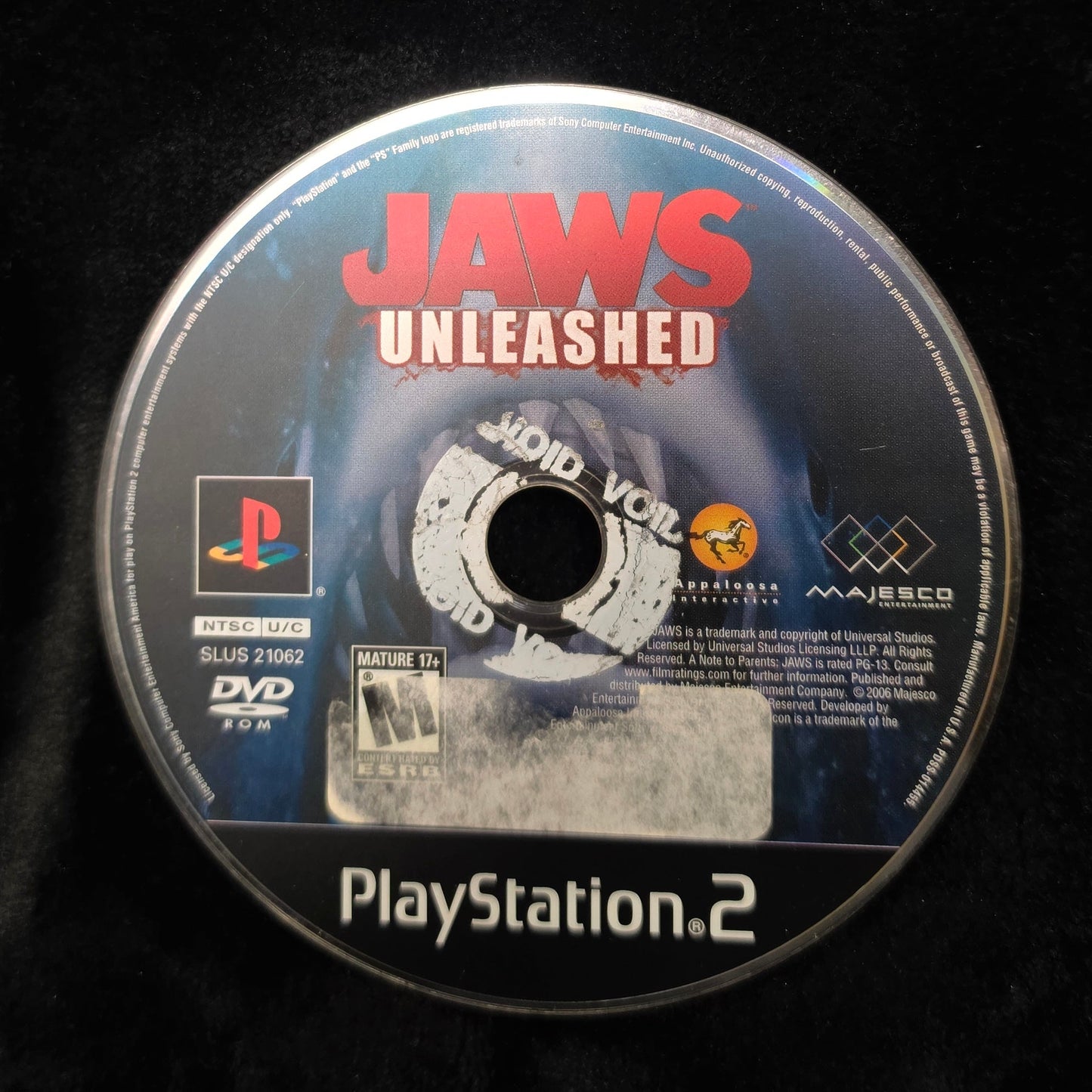 A PlayStation 2 game disc for Jaws Unleashed resting on a black fabric surface, with a PlayStation logo and PlayStation 2 branding visible on the disc.