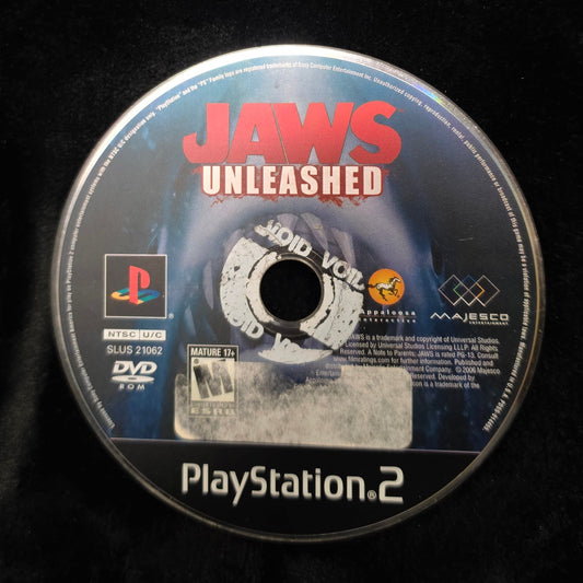 A PlayStation 2 game disc for Jaws Unleashed resting on a black fabric surface, with a PlayStation logo and PlayStation 2 branding visible on the disc.