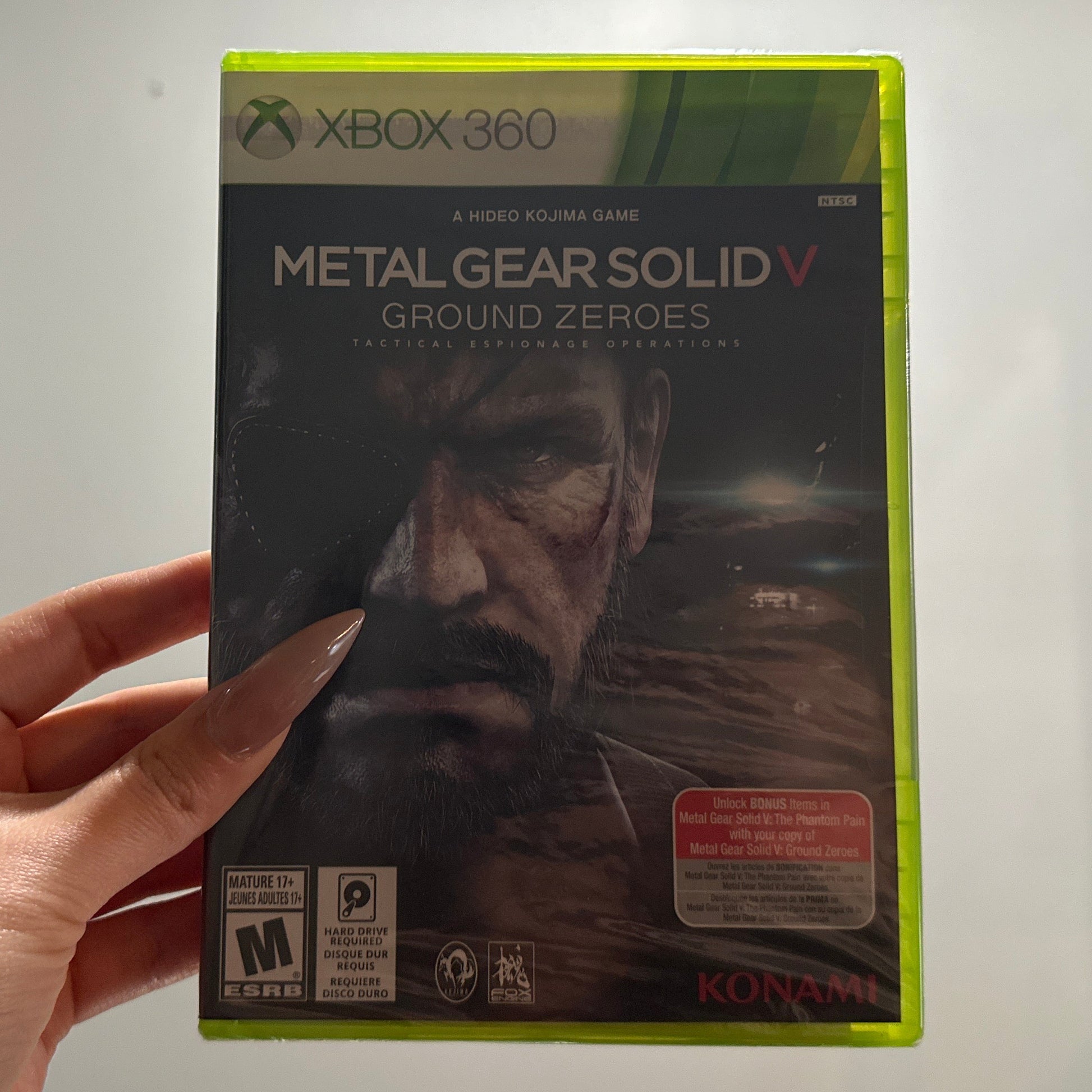 Hand holding a 'Metal Gear Solid V' game box for Xbox 360 against a neutral background