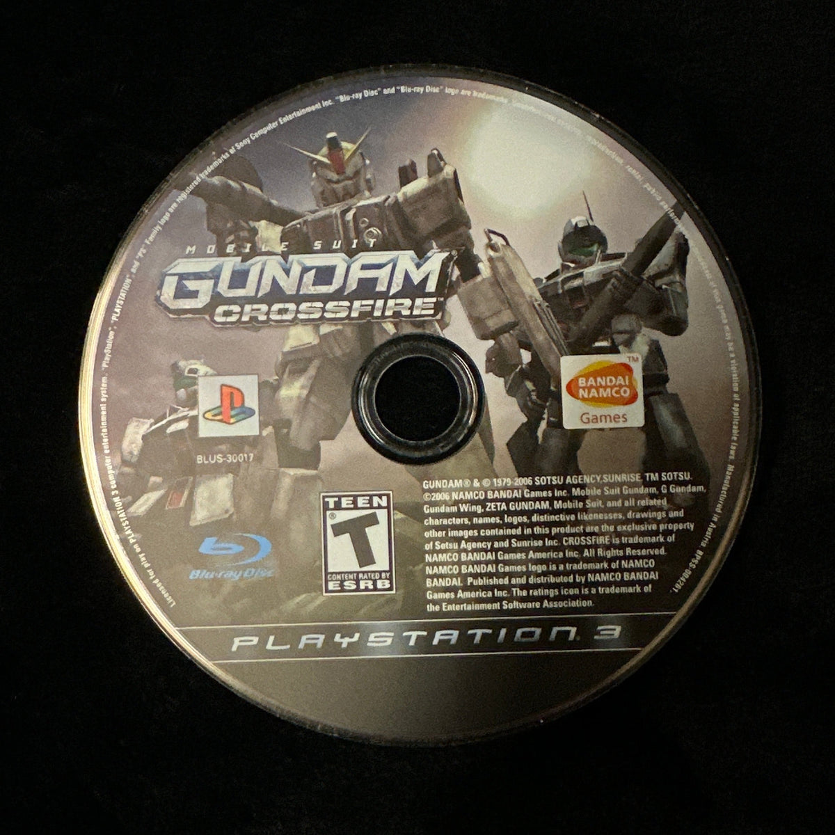 A PlayStation 3 game disc for Gundam Crossfire resting on a black fabric surface.