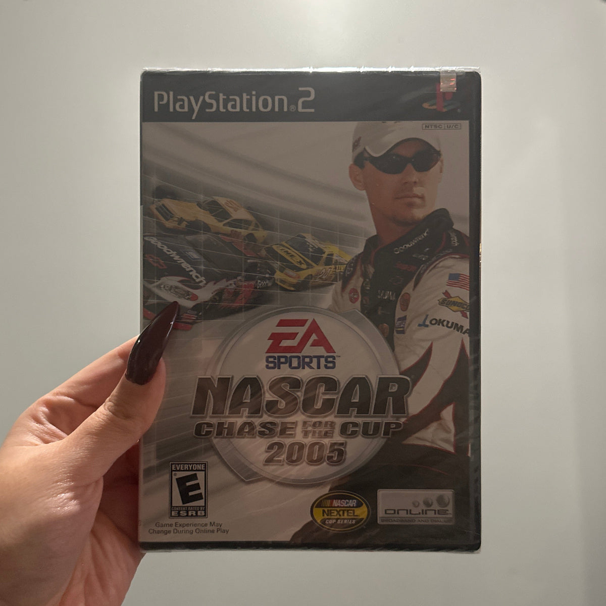 A hand holds a PlayStation 2 game case for EA Sports NASCAR Chase Cup 2005 against a plain indoor wall.