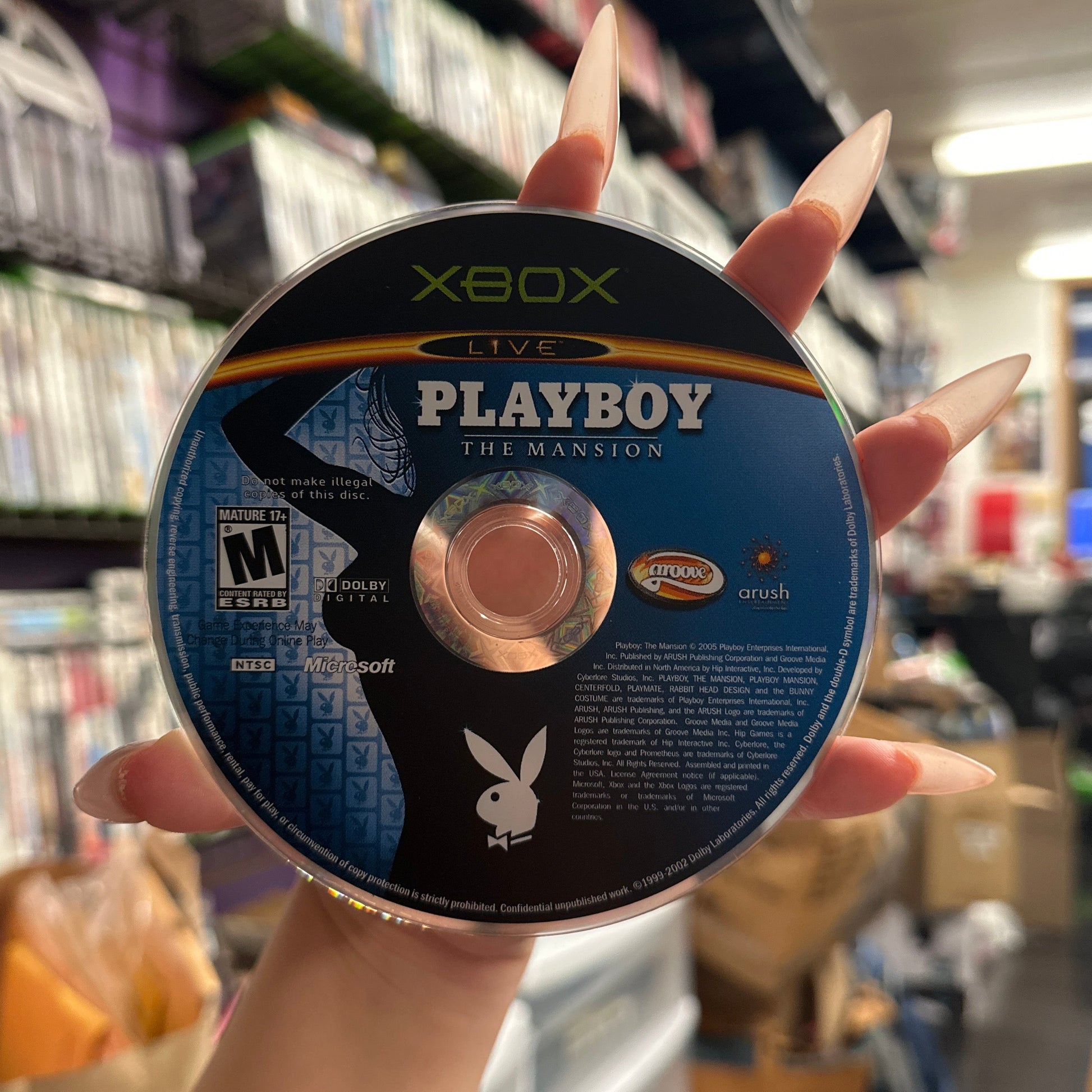 A hand with long manicured nails holds a Xbox Live Playboy The Mansion game disc in a retail store setting with shelves of video games blurred in the background.
