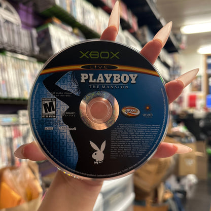 A hand with long manicured nails holds a Xbox Live Playboy The Mansion game disc in a retail store setting with shelves of video games blurred in the background.