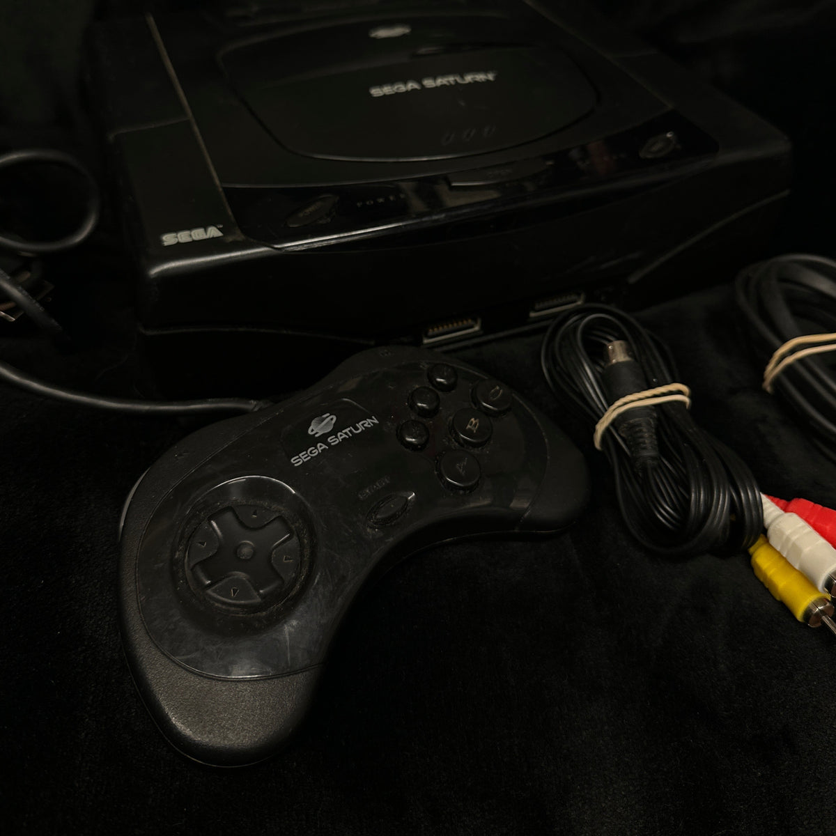 A black Sega Saturn console with a matching wired controller, power cable, and red/white/yellow audio cables, displayed on a black fabric surface.