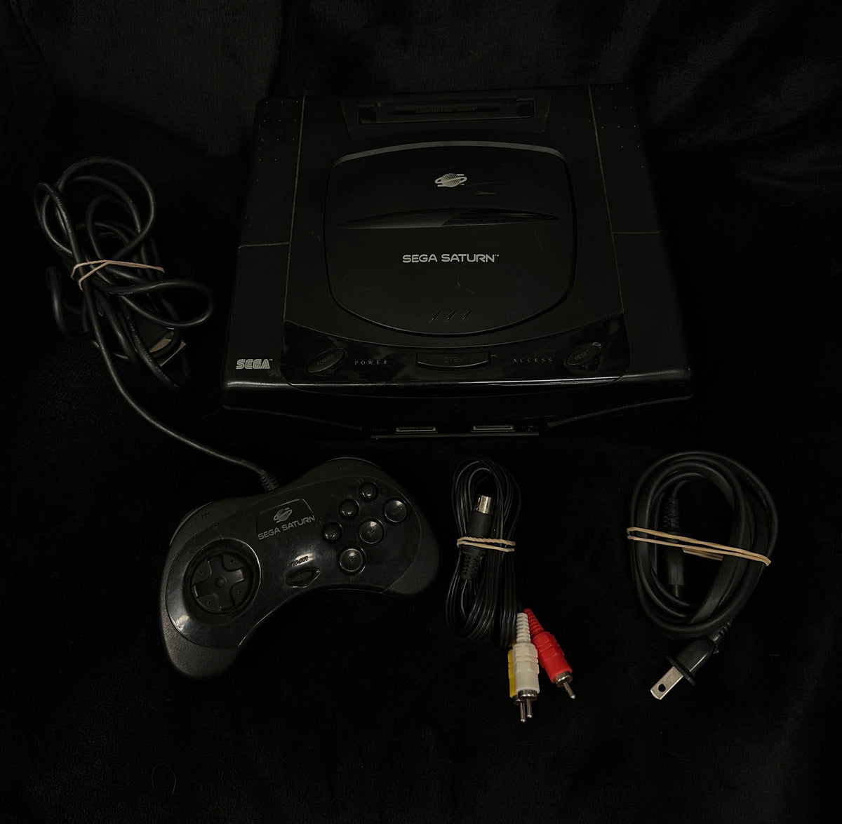 A black Sega Saturn console with its original controller, power cable, and audio/video cables laid out on a black fabric background.