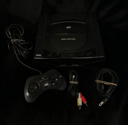 A black Sega Saturn console with its original controller, power cable, and audio/video cables laid out on a black fabric background.
