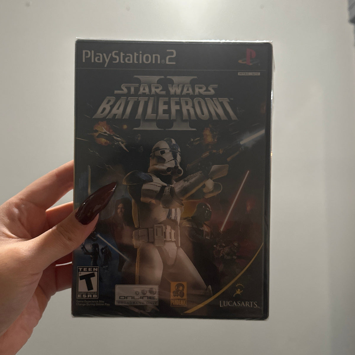 A hand holding a PlayStation 2 game case for Star Wars: Battlefront II against a plain indoor wall.