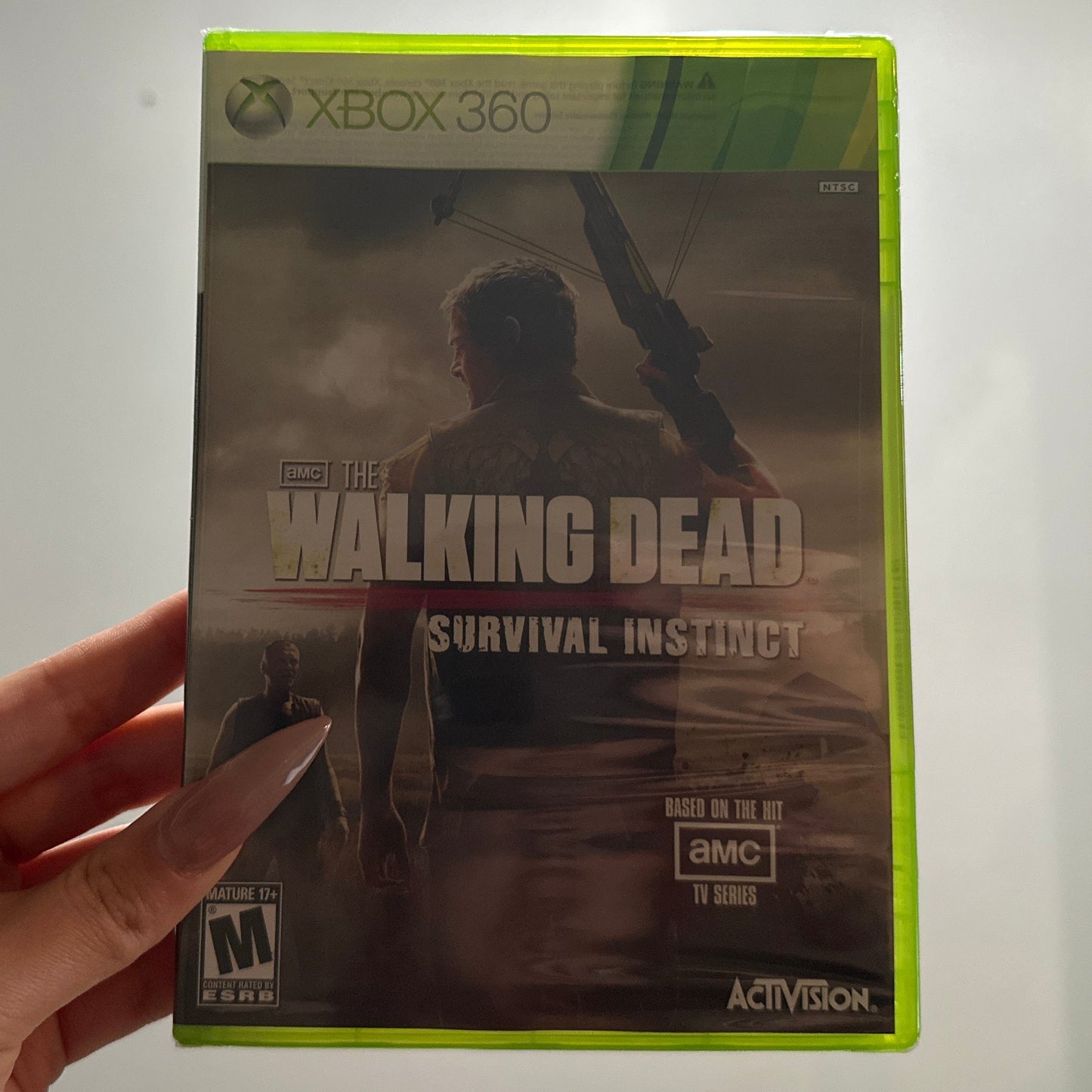 Hand holding a 'The Walking Dead: Survival Instinct' Xbox 360 game case against a neutral background