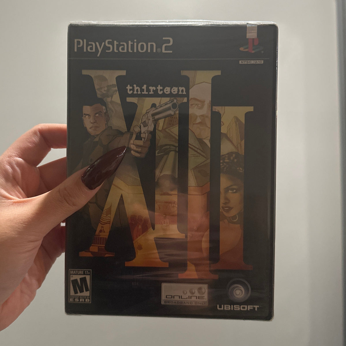 A hand with dark red nail polish holds a PlayStation 2 game case for 'Thirteen' by Ubisoft. The game cover features a dark design with large Roman numerals and character artwork, set against a plain indoor wall background.