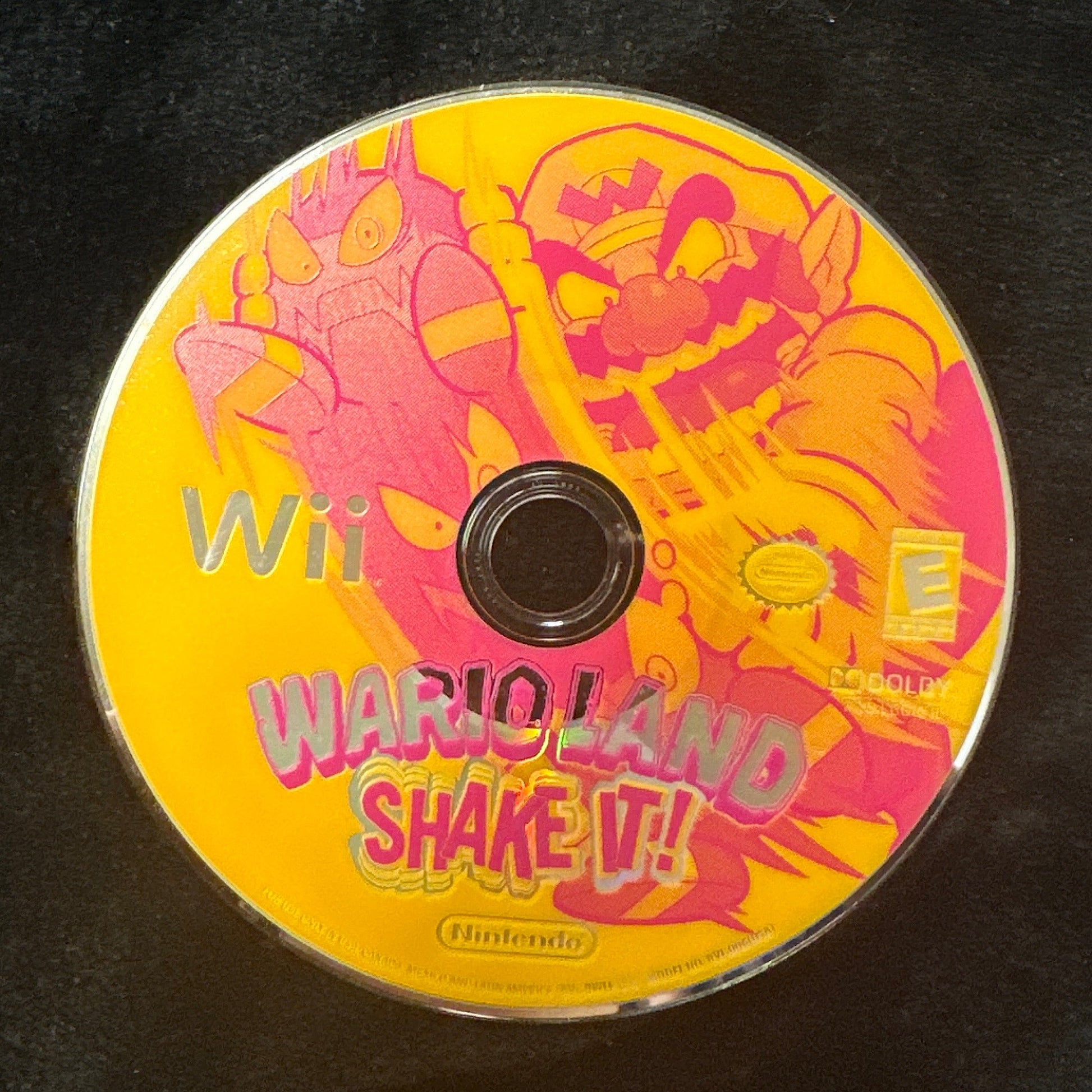 A Wii game disc for 'Wario Land Shake It!' resting on a black fabric surface, with a dark tiled floor visible in the background.